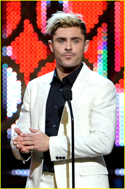 Zac Efron has blonde hair and wears a white suit jacket on stage at an awards show